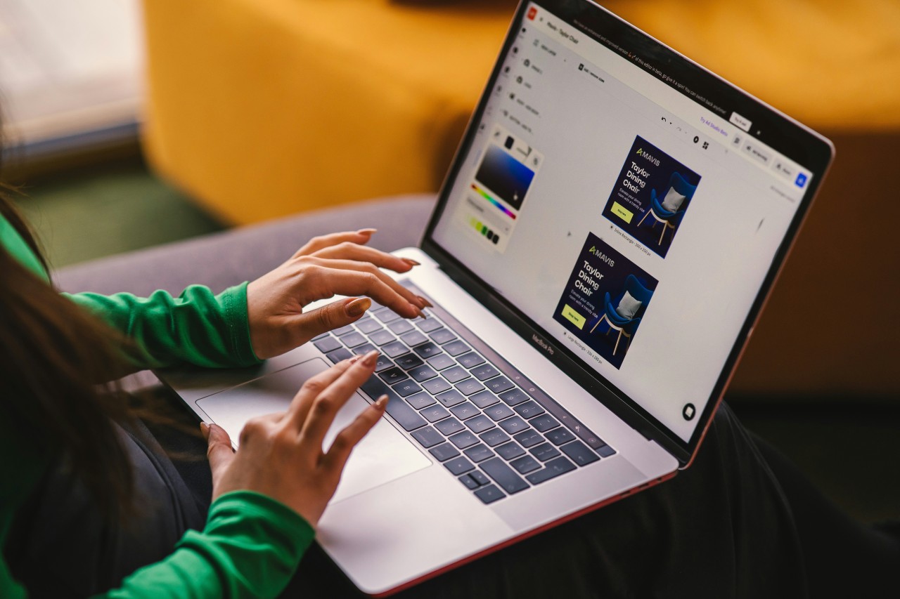 A woman in a green long-sleeve shirt working on a MacBook Pro laptop displaying a graphic design tool with advertising banner templates on screen